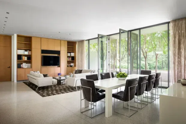 A bright, spacious open-plan living and dining area with floor-to-ceiling glazed folding or sliding doors opening to greenery. The room features extensive warm timber built-in cabinetry with integrated shelving and television joinery, a neutral seating area, a white dining table with dark leather chairs, a pale tiled or polished floor, and minimal recessed lighting. The overall design is clean, refined, and contemporary with strong indoor-outdoor connection.