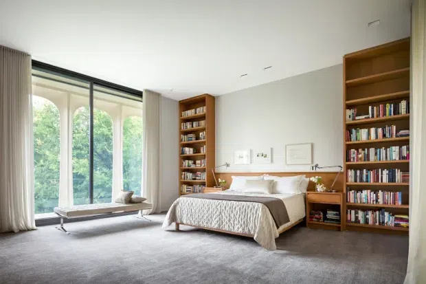 A spacious, refined bedroom with a large upholstered bed positioned between two built-in timber bookcases. Full-height glazed doors or windows with sheer curtains open to leafy views, while a low upholstered bench sits by the glazing. The room has a pale neutral palette, grey carpet, warm timber joinery, restrained artwork and soft natural light.