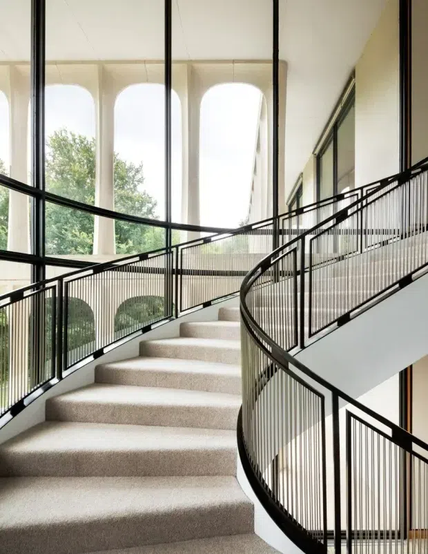 A bright, elegant interior stairway with broad carpeted steps curving upward through a multi-level landing. Slim black metal balustrades with closely spaced vertical pickets define the sweeping staircase and overlook. Tall glazed openings and large arched exterior openings bring in abundant natural light, with trees visible beyond. The space has a refined, contemporary architectural character and a high, airy volume.