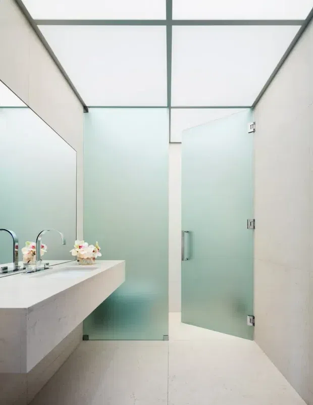 Bright contemporary bathroom with a floating pale stone vanity, twin chrome faucets and a large wall mirror. Frosted glass shower screens and door create a translucent enclosure, while a gridded translucent ceiling and light neutral surfaces give the space an airy, minimalist character.