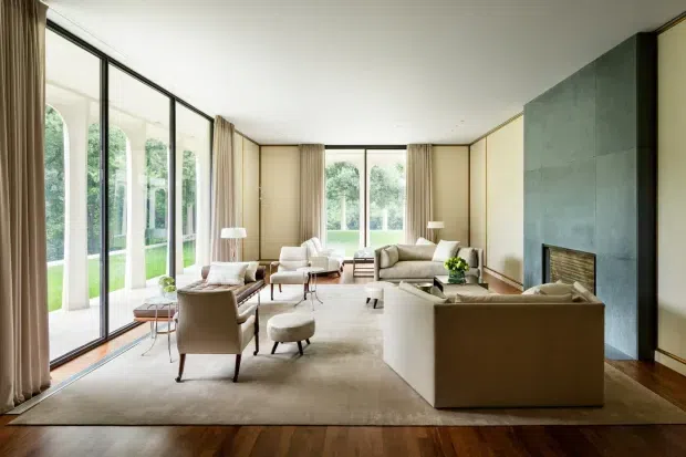 A refined, spacious living room with floor-to-ceiling glazed doors and windows along one side, opening visually onto a green garden. The room features a large neutral rug, several cream and taupe upholstered sofas and armchairs, small side tables, lamps, and a prominent dark green-grey stone fireplace wall with a built-in fireplace. Warm timber flooring and beige curtains create a calm, sophisticated atmosphere.