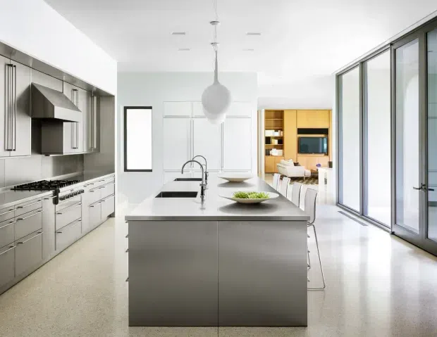 Bright, spacious contemporary kitchen with a large central island, integrated sink and seating, extensive white cabinetry, stainless-steel appliances, and a grey-toned backsplash. A dining and living area is visible beyond the kitchen, with full-height glazed doors along the right side bringing in abundant natural light. The space has clean lines, minimalist detailing, and a neutral monochromatic palette.