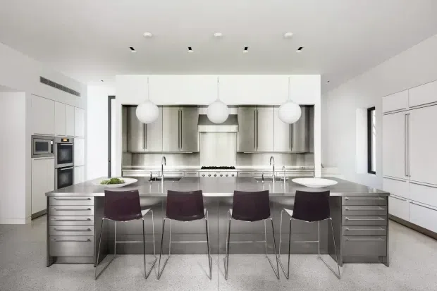 A bright, spacious contemporary kitchen with a large central island, four bar stools, extensive stainless-steel and white cabinetry, integrated appliances, twin sinks, and a professional-style cooking area. Three spherical pendant lights hang above the island, with recessed ceiling lighting and a light terrazzo-like floor creating a clean, refined appearance.