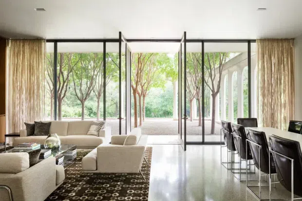 A bright, open-plan contemporary interior combining a living room and dining area. Floor-to-ceiling black-framed glazing and central open glass doors connect the room to a landscaped courtyard. The living area contains cream sofas and armchairs arranged around a dark patterned rug, while a long dark dining table with high-back chairs occupies the right side. Neutral sheer curtains, polished light flooring, and a clean white ceiling create an airy, refined atmosphere.