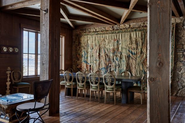 Rustic dining area in a timber-framed interior, featuring a long dark dining table with ornate upholstered chairs, wide plank timber flooring, exposed structural beams and rafters, dark timber wall panelling, and a rough stone feature wall. A large textured woven wall hanging forms a focal point behind the table, while tall windows provide views of a bright coastal landscape.