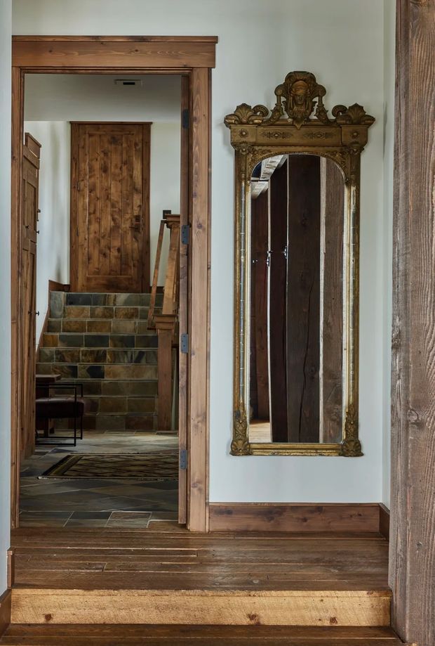 Interior view of a rustic traditional hallway or landing with a broad timber-framed doorway, dark reclaimed wood flooring and trim, and a richly carved antique gold-framed wall mirror. Through the doorway are stone-tiled steps, timber doors, and additional rustic wood detailing. The space has a warm, heritage-inspired character with natural timber, muted pale walls, and mixed stone flooring.