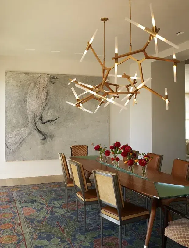 A refined dining room with a long dark timber dining table surrounded by upholstered dining chairs. A sculptural branching chandelier with warm brass-toned arms and linear white lights hangs prominently above the table. The room features a large monochromatic abstract artwork, patterned area rug, white walls, and floral arrangements on the table. The overall look combines contemporary lighting with mid-century-inspired furniture and layered decorative detailing.