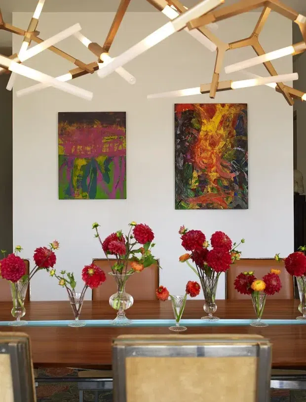 A bright, contemporary dining room viewed toward a white feature wall with two colourful abstract artworks. A long dark timber dining table is arranged with numerous clear glass vases of red, pink and orange flowers, while a sculptural suspended ceiling light made from angular timber arms and white linear tubes dominates the ceiling. A dining chair is visible in the foreground.