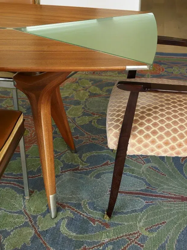 Close-up of a mid-century dining or meeting table with warm wood surfaces and tapered legs, paired with a dark wood-framed upholstered chair. A distinctive pale green translucent curved glass or acrylic table extension runs along the tabletop edge. The furniture sits on a patterned multicolour carpet in an interior setting.