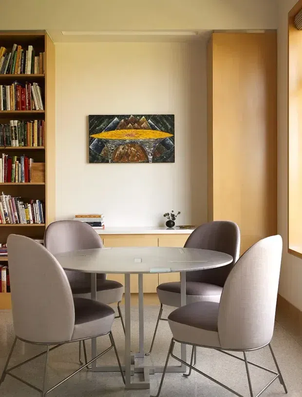 A warm, refined dining area with a round pale table and four upholstered dining chairs. Full-height built-in timber shelving filled with books occupies the left wall, while pale timber cabinetry and a large timber-framed window line the room. A small abstract artwork hangs above the cabinetry, and terrazzo-like flooring completes the space.