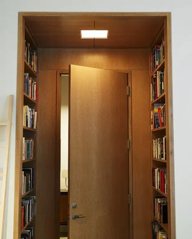 A narrow built-in timber-lined passage or doorway framed by full-height bookshelves on both sides. A solid timber door is partly open, revealing a small adjoining room beyond. The ceiling and walls are clad in warm medium-brown wood, with a small rectangular recessed ceiling light and integrated shelving filled with books.