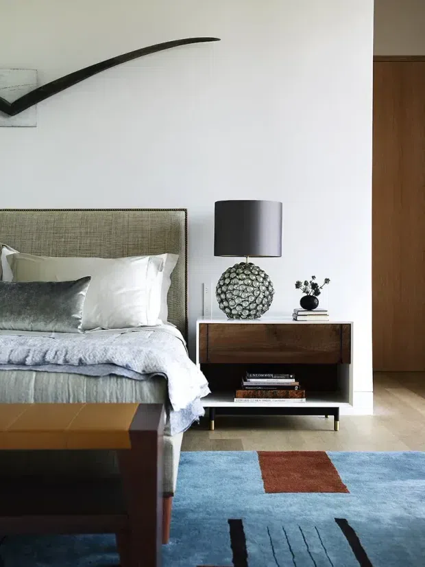 A refined bedroom interior featuring a partially visible upholstered bed with textured grey headboard, white bedding, and a dark accent cushion. A mid-century-inspired timber bedside cabinet with open shelving sits beside the bed, topped with a sculptural spherical lamp and small black vase. White walls, timber flooring, a colourful geometric rug, and a dark abstract wall sculpture complete the room.