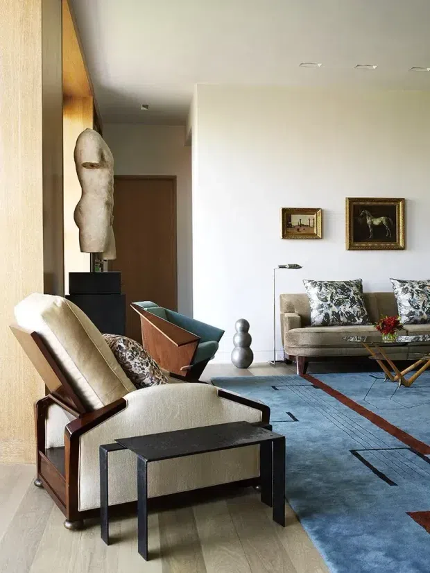 A refined living room with a mid-century-inspired lounge chair and ottoman in the foreground, a sculptural black pedestal, a textured abstract sculpture, and a vintage-style sofa arranged around a patterned blue rug. The room has warm timber wall panelling and joinery, pale walls, recessed ceiling lights, framed artwork, and a mix of neutral upholstery with dark wood and black accents.