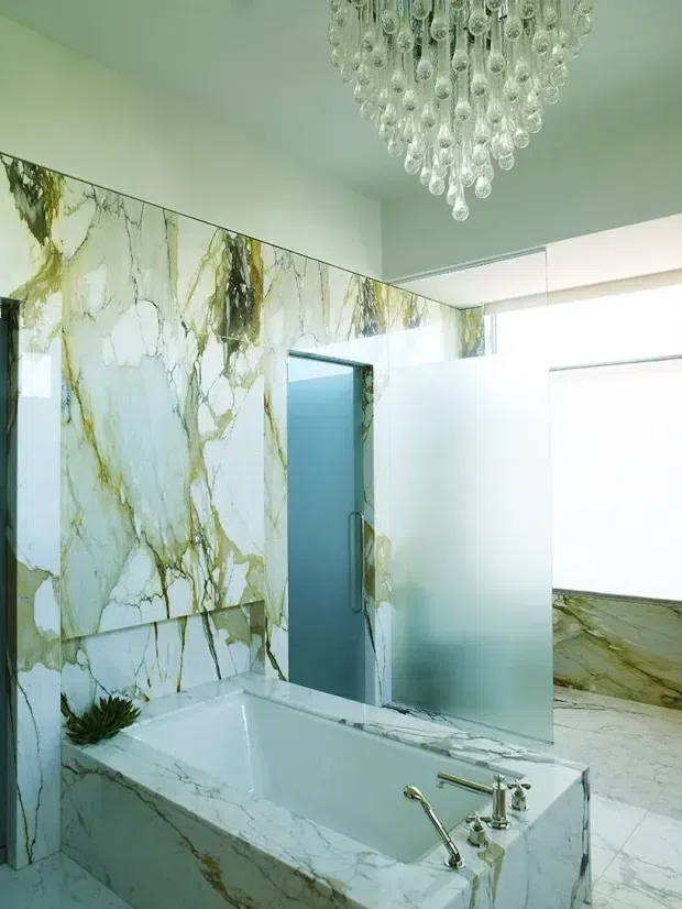 Luxurious contemporary bathroom featuring a freestanding-style rectangular bathtub enclosed in a dramatic veined white-and-green marble surround, matching full-height marble wall panels and flooring, a frosted glass shower or bathroom partition, and a large crystal drop chandelier. The space is bright and refined with strong natural stone detailing.