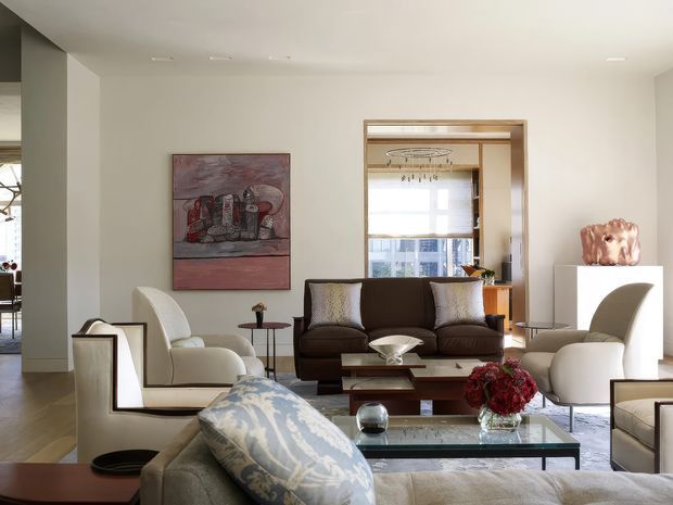 A bright, refined living room with a contemporary-classic composition. The space features a large neutral sectional sofa in the foreground, a dark brown leather sofa, sculptural cream armchairs, a glass coffee table, and a timber coffee table. A timber-framed glazed built-in shelving or doorway is positioned behind the seating, with recessed ceiling lighting, pale walls, timber flooring, artwork, and carefully styled decorative objects. The room opens toward a dining area at the left and has an airy, curated atmosphere.