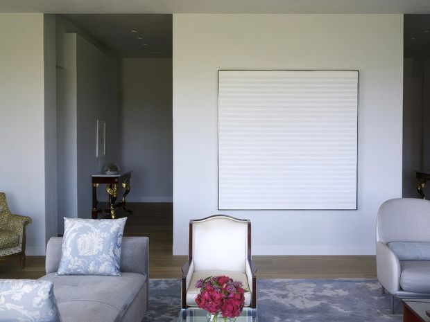 A refined contemporary living room with pale grey upholstered seating arranged around a central coffee table with flowers. A large framed monochrome textured artwork hangs on a light grey feature wall, while a darker hallway or entry zone with a small console table is visible to the left. The room has a muted, elegant palette and a patterned area rug.