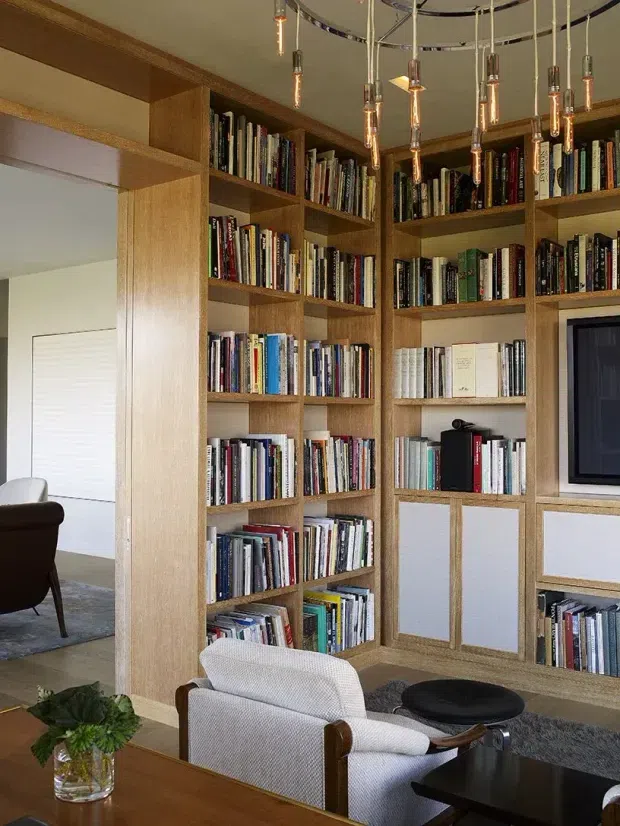 Interior view of a refined library and living area dominated by custom floor-to-ceiling timber shelving filled with books. The built-in joinery incorporates lower closed cabinets and a television niche. A sculptural cluster of hanging pendant lights, upholstered lounge chairs, a coffee table, rug, and small plant complete the warm, curated space.