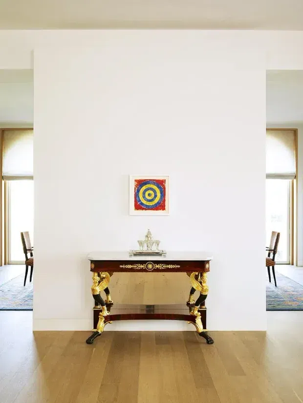 A bright, symmetrical interior vignette featuring an ornate console table centred against a tall white wall. The console has dark wood, gilded sculptural legs and a reflective stone or mirrored apron, with small decorative objects on top. A colourful concentric-circle artwork hangs above it. Openings on both sides reveal adjacent spaces with dining chairs, patterned rugs and tall windows. The room has pale timber flooring, white walls and a light neutral ceiling.