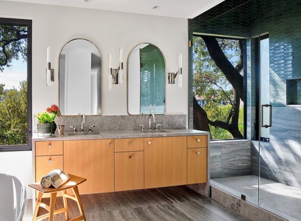 Bright, refined bathroom featuring a long floating timber double vanity with twin undermount sinks, grey stone benchtop, arched mirrors, wall sconces, and a frameless glass shower enclosure. The shower has grey stone-look wall and floor finishes with a dark green textured tile feature ceiling or upper wall. Large windows overlook leafy trees, bringing in natural light.