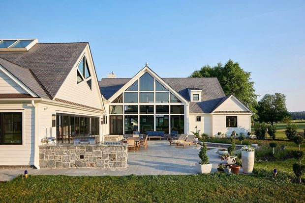 Exterior view of a large, light-coloured residential house with multiple gabled volumes, dark shingle roofs, prominent floor-to-ceiling glazed gable frontage, and a spacious paved patio with outdoor seating. The home sits in a landscaped rural or semi-rural setting.
