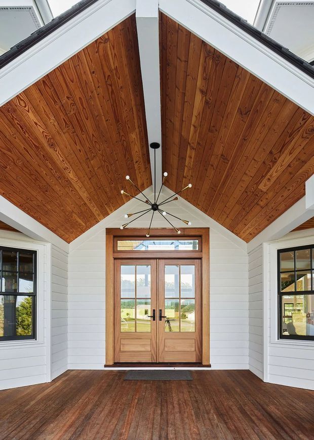 A bright, symmetrical entry porch or foyer with a dramatic vaulted ceiling clad in warm timber boards. White horizontal wall cladding and structural beams frame a central timber double door with glazed panels and a transom window. Matching black-framed windows flank the entrance, while a sculptural multi-arm pendant light hangs from the ceiling. The floor is finished in dark timber boards.