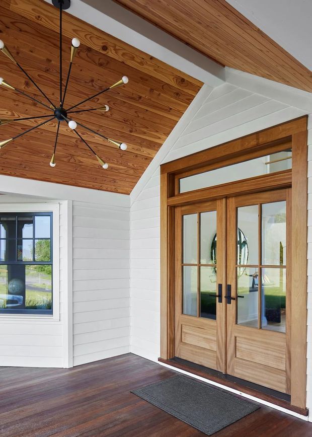 A bright covered entry porch featuring white horizontal weatherboard walls, a warm timber-lined vaulted ceiling, dark timber flooring, and a substantial timber-framed glazed double front door with a transom window. A black multi-arm pendant light hangs from the ceiling, and a side window overlooks the grounds.