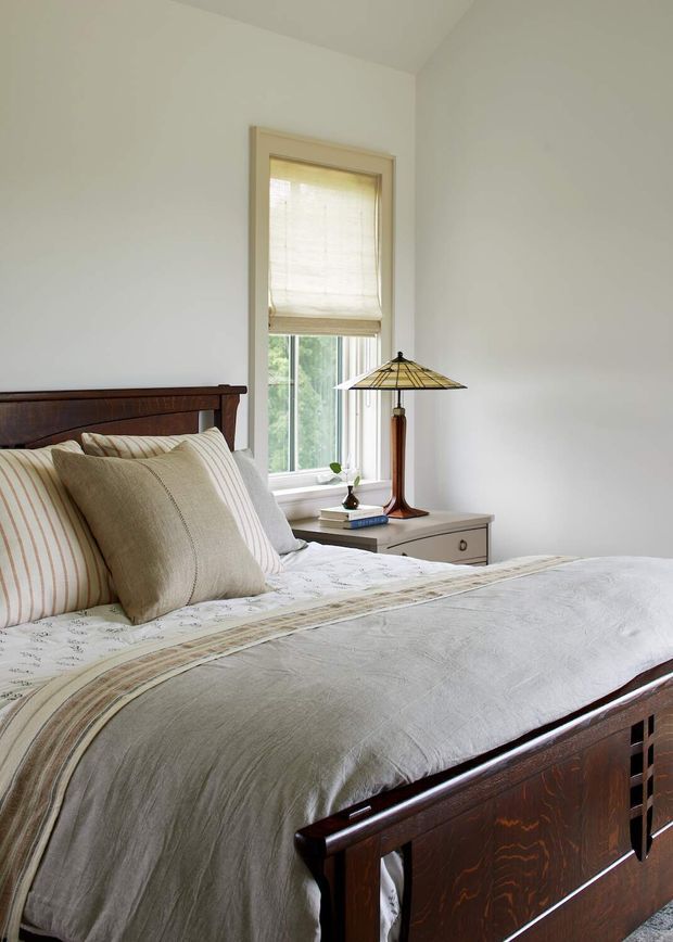 A calm, traditionally styled bedroom with a dark stained timber bed, cream and beige striped bedding, a light wood bedside table, and a classic stained-glass table lamp. A tall window with a woven Roman blind brings in soft natural light, while the walls and ceiling are painted off-white.