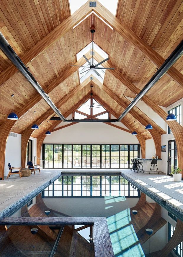 A spacious indoor swimming pool housed beneath a dramatic vaulted timber ceiling. Exposed wood paneling and structural beams frame the room, with exposed metal ductwork, ceiling fans, blue pendant lights, and extensive black-framed glazing. A bar-height counter and stools occupy one side, while the pool reflects the large windows and ceiling structure.