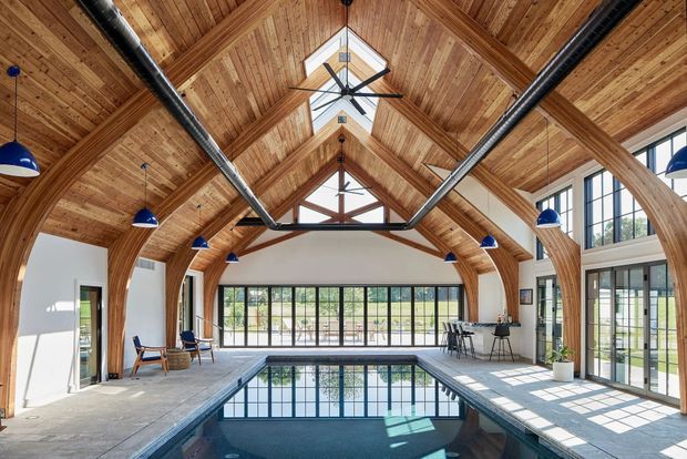 A bright indoor swimming pool housed in a dramatic timber-vaulted space. The room features exposed arched timber framing and timber ceiling boards, extensive black-framed glazing, skylights, exposed dark metal ductwork, blue pendant lights, and a tiled pool surround. Large windows overlook greenery outside.