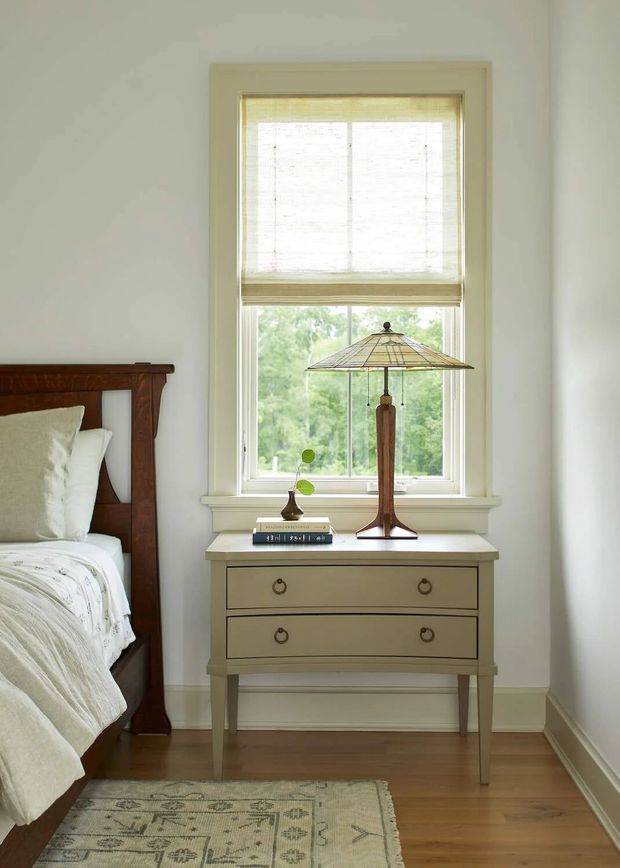 A bright, traditional bedroom corner featuring a dark timber bed, pale upholstered bedding, and a painted two-drawer bedside chest beneath a tall window. The window has light woven Roman blinds, and a stained-glass-style table lamp with a timber base sits beside a small vase and books. Hardwood flooring, white walls and trim, and a muted patterned rug create a calm, warm interior.