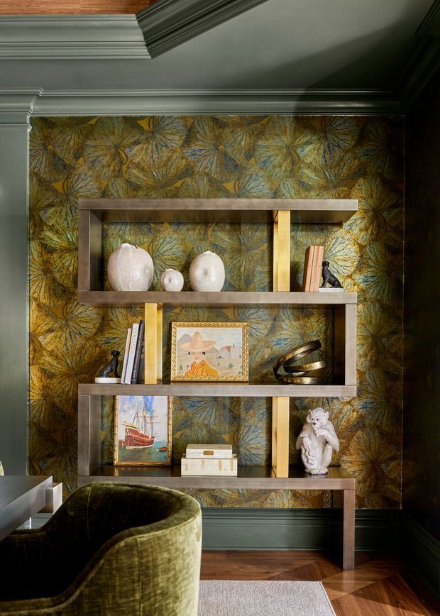 A richly layered interior vignette featuring a dark wood open display shelving unit against botanical-patterned wallpaper. The shelves hold ceramics, books, framed artwork, a sculptural object, a storage box and a decorative figurine. Deep green painted walls and ornate ceiling cornices frame the composition, while a plush olive-green upholstered chair, timber flooring and a pale rug are visible in the foreground. The overall look is warm, dramatic and refined.