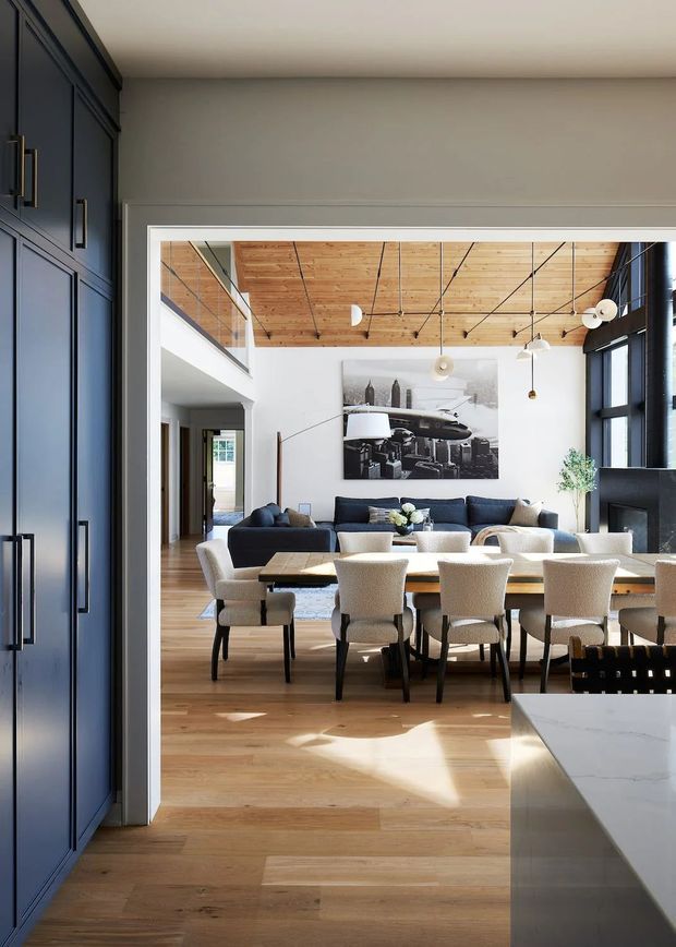A refined open-plan interior viewed from a dark navy built-in cabinetry zone, looking toward a dining and living area. The room features wide-plank timber flooring, a timber-paneled ceiling with exposed dark linear detailing, large black-framed windows, a long dining table with upholstered cream chairs, dark blue sofas, sculptural pendant lights, and a monochrome artwork. A pale stone kitchen island is visible in the foreground, with a double-height opening and mezzanine-like upper-level void beyond.
