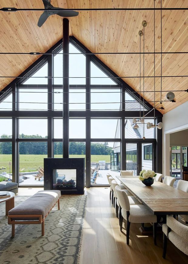 A bright, open-plan dining and living space with a dramatic double-height glazed gable wall, exposed timber vaulted ceiling, dark structural framing, and a central black fireplace. A large timber dining table with upholstered chairs occupies the right side, while a sofa and bench sit on a patterned rug to the left. Floor-to-ceiling glazing opens toward a sunny rural landscape and outdoor terrace.