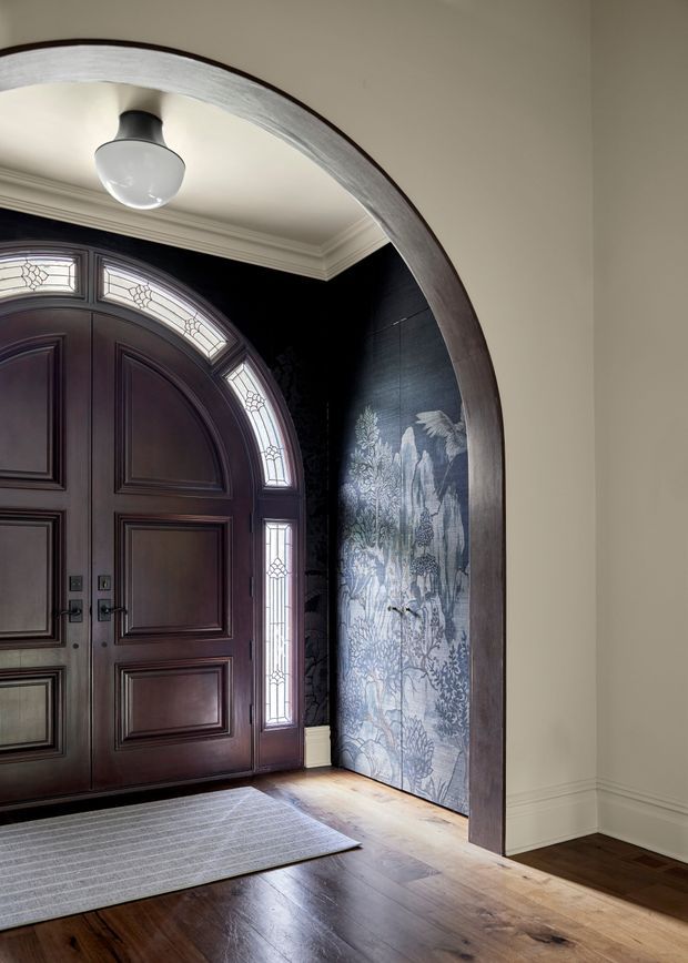 A refined residential entryway viewed through a large arched opening. The space features a dark stained, panelled double front door with decorative leaded-glass sidelights and fanlight, a dramatic dark botanical mural or wallpaper on the adjacent wall, white decorative ceiling moulding, warm timber flooring, and a simple pendant light. The overall composition is elegant and traditional with a high-contrast, moody palette.