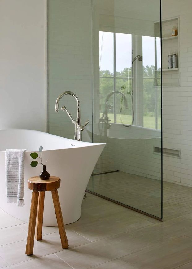 Bright contemporary bathroom featuring a sculptural freestanding white bathtub, large frameless glass shower enclosure, wall-mounted open shelving, brushed metal tapware, white brick-pattern wall tiles, and pale large-format floor tiles. A small timber stool with a vase and towel adds natural warmth.