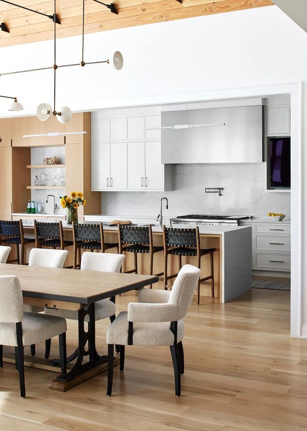 A bright open-plan kitchen and dining area with a warm timber ceiling, light timber flooring, white built-in cabinetry, a pale stone backsplash and island, and dark-framed dining and bar furniture. The kitchen includes a long island with seating, integrated appliances, open timber shelving, pendant lighting, and a sculptural dining table with upholstered chairs in the foreground.