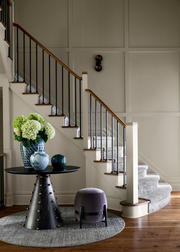 A refined residential stairway and entry landing with a curved staircase, white paneled walls, dark timber handrails, slim black balusters, and patterned carpet runners. A round black console table with decorative vases and a small upholstered stool sits on a textured circular rug beside the stairs. Warm timber flooring and soft neutral finishes create an elegant, welcoming atmosphere.