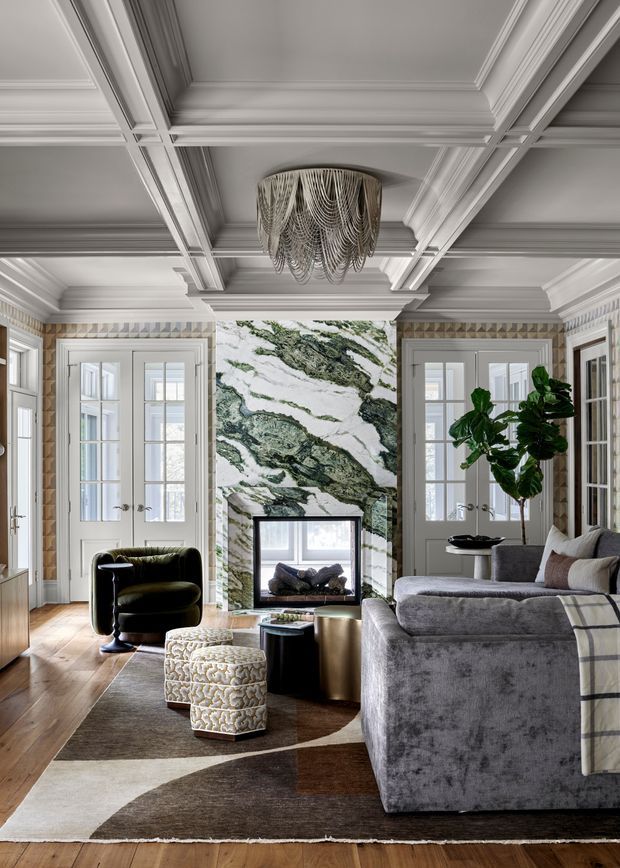 Elegant, richly detailed living room with a coffered and moulded white ceiling, timber flooring, French doors, patterned wall detailing, and a dramatic green-and-white marble fireplace surround. The room is furnished with a large grey sectional sofa, dark accent chair, sculptural upholstered ottomans, metallic side tables, a statement chandelier, and a large area rug. The styling combines classic architectural detailing with contemporary, luxurious furnishings.