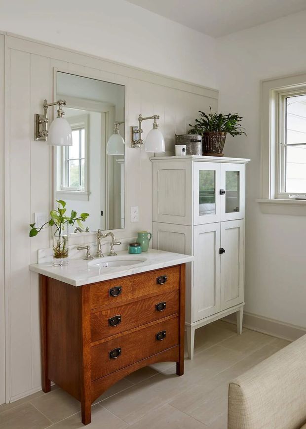 A bright, traditional bathroom featuring a freestanding timber vanity with a white integrated basin and polished metal tapware, a large wall mirror, paired wall-mounted lights, white vertical wall panelling, and a tall painted storage cabinet with glazed doors. A window provides natural light, while potted plants and a partial upholstered seat add warmth.