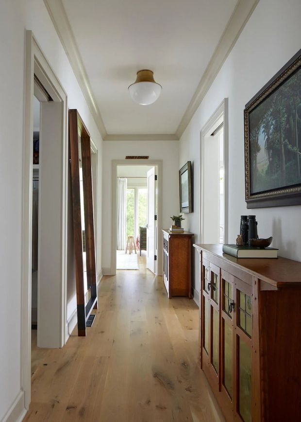 A bright, narrow residential hallway with wide-plank light timber flooring, white walls, decorative ceiling cornice, and a central flush-mount globe light. Timber sideboards and cabinetry line the right wall, while framed artwork, a tall mirror, and doorways lead toward a sunlit room at the end.