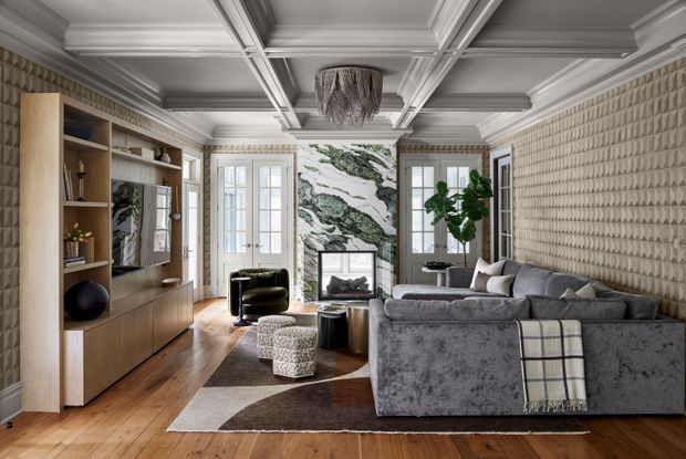 Sophisticated living room with a large grey sectional sofa, built-in timber media and shelving unit, marble fireplace surround, patterned feature wall, coffered ceiling with detailed moulding, hardwood flooring, and elegant layered lighting. Tall French doors bring in natural light, while sculptural furniture and a geometric rug create a refined contemporary-classic atmosphere.