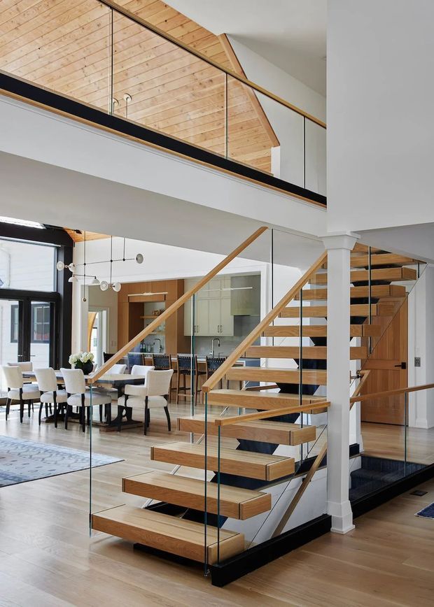 A bright, contemporary open-plan interior centred on a floating timber staircase with clear glass balustrades, slim dark metal supports, and timber handrails. The stair rises beside a white structural column to a mezzanine or upper landing with timber-clad angled surfaces and glass guarding. A dining area, kitchen, timber cabinetry, large windows, and polished light timber flooring are visible in the background. The design combines warm natural timber with white walls, black accents, and abundant daylight.