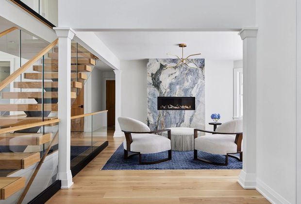 A refined contemporary living room viewed from an open stairway, featuring a sculptural marble-look fireplace surround with linear firebox, two curved upholstered lounge chairs, a dark blue area rug, a low textured coffee table, and a dramatic modern chandelier. The adjacent staircase has warm timber treads, slender timber handrails, and clear glass balustrades, while white columns, trim, and walls create a bright, polished interior.