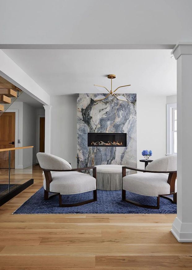 A refined living room with a central marble fireplace surround and built-in linear fire, two upholstered mid-century-inspired lounge chairs, a round ribbed ottoman, and a dark blue patterned rug. The room features white walls and ceiling, warm timber flooring, classic column and trim details, a sculptural brass pendant light, and a glimpse of adjoining cabinetry and hallway.