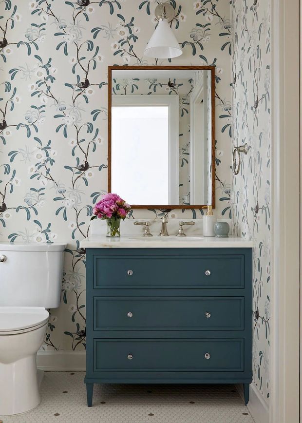 A compact, traditionally styled bathroom featuring a blue three-drawer vanity with a white stone countertop, chrome fixtures, a framed wall mirror, a white toilet, and patterned botanical wallpaper. The floor is finished in small white hexagonal tiles with dark accents, and a wall-mounted pendant adds a decorative focal point.