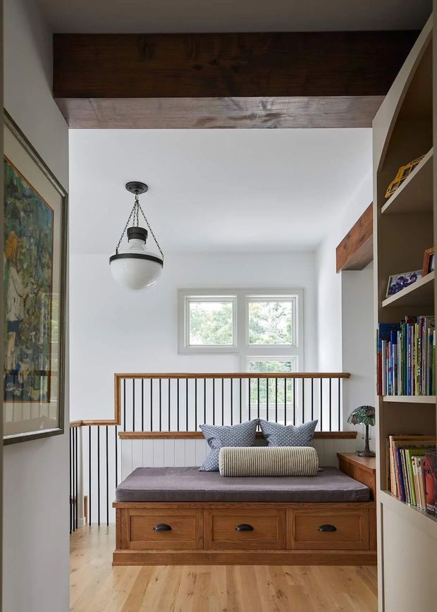 Bright, compact landing or reading nook with a built-in timber window seat beneath a multi-pane window, fitted drawers, cushions, and surrounding open bookshelves. A black metal balustrade overlooks the level below, while exposed dark timber beams and a hanging pendant add architectural character. The space has white walls, pale timber flooring, and warm natural finishes.