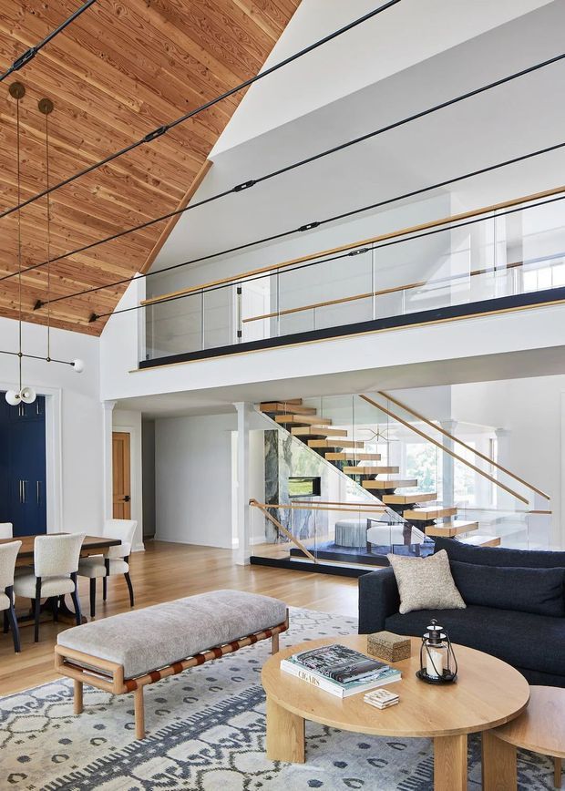 A bright, open-plan contemporary living and dining area with a dramatic double-height volume. The room features a warm timber-lined pitched ceiling, white walls, a floating open-riser staircase with glass balustrades, a mezzanine level, and slim black structural or lighting elements. A dark navy sofa, round timber coffee table, upholstered bench, dining table and patterned rug furnish the space.