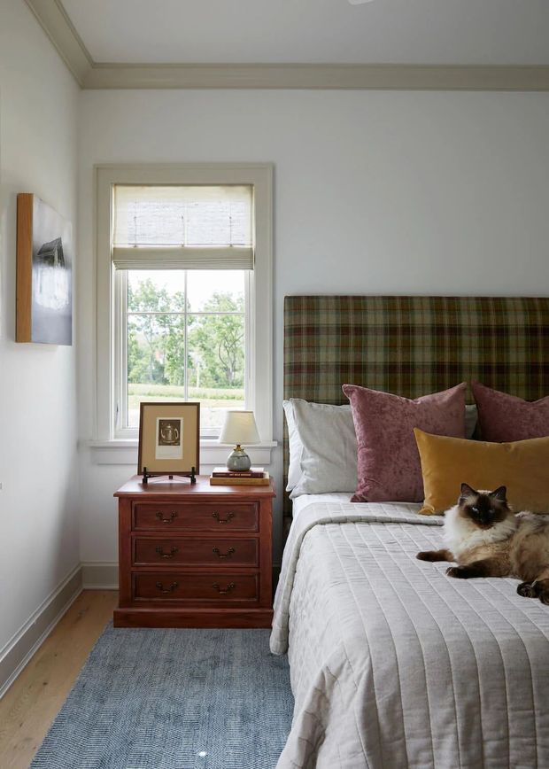 A warmly styled bedroom with a traditional upholstered plaid headboard, white bedding, mauve and mustard cushions, and a dark wood three-drawer bedside chest. A tall sash window with a Roman blind brings in natural light, while a blue-grey rug and framed artwork add subdued colour. A cat is resting on the bed.