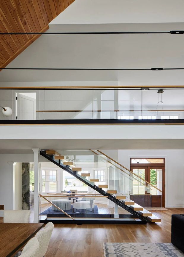 Interior view of a bright, multi-level living area centred on a contemporary open-riser staircase with black structural stringers, timber treads, and clear glass balustrades. White walls, exposed timber ceiling lining, polished timber flooring, large windows and glazed doors create an airy open-plan space. A mezzanine or upper-level walkway is visible above, with linear black pendant lighting spanning the ceiling.