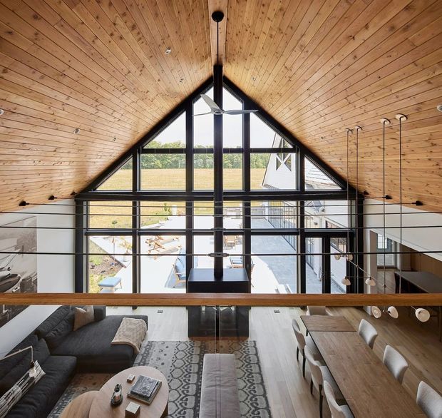 A dramatic double-height interior viewed from an upper-level landing or mezzanine, featuring a steep timber-lined pitched ceiling, exposed black structural framing, and expansive black-framed glazing at the gable end. The lower level contains an open-plan living and dining area with a dark sectional sofa, timber dining table, and pendant lights, while a central suspended fireplace or chimney element and simple cable balustrade define the mezzanine edge.